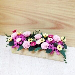 Miniature pink and yellow flower arrangement with leaves in a wooden planter on a light surface.