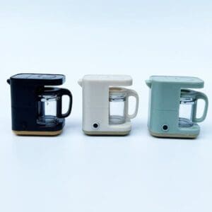 Three small coffee makers in black, cream, and mint green are arranged in a row on a light background.