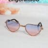 Heart-shaped sunglasses with gradient pink-blue lenses, black arms, and gold frame on a soft pastel background.
