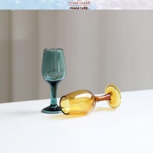 Two miniature wine glasses, one upright in teal and one lying down in amber, on a white surface.