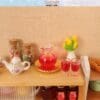 Miniature kitchen scene with a tea set, colorful candy jars, a jug of red drink, two glasses, and yellow tulips in a vase.