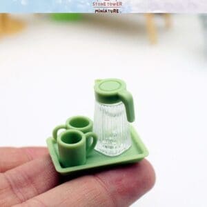 A tiny green tray holds a clear pitcher and two green mugs on a person's fingertip.