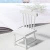 Miniature white chair placed on an open book, with soft lighting and a blurred plant in the background.