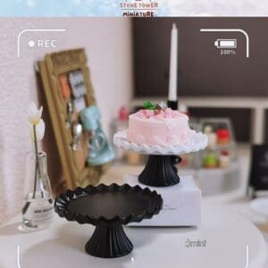 Miniature pink cake on a white stand, beside a black stand, with a small vase on a table in a cozy setting.