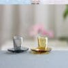 Two miniature glass cups with saucers, one clear and one amber, on a textured surface.