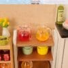 Miniature kitchen setup with colorful juice pitchers, cooking pots, jam jars, and a vase of yellow flowers on wooden shelves.