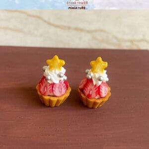Two miniature strawberry tarts with cream, silver balls, and yellow star toppers displayed on a wooden surface.