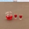 Miniature clear jug and two small glasses filled with red liquid on a brown surface.