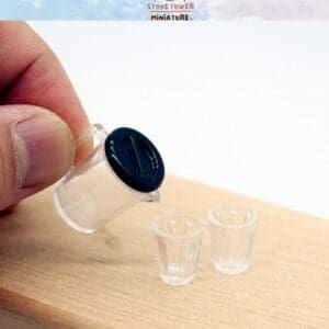 Hand holding mini pitcher, pouring into two tiny clear glasses on a wooden surface.