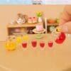 Miniature kitchen scene with tiny teacups, pastries, and colorful drinks on a table; hand pouring red juice from a small pitcher.