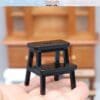 Miniature black step stool held in a hand with a wooden cabinet in the background.