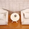 Two white armchairs with pillows facing a round table holding a bubble tea against a tiled wall on a wooden floor.