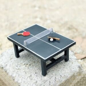 Miniature ping pong table with a red and a black paddle, and a ball, on a concrete surface.