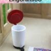 Miniature white trash bin with red lid and foot pedal on a table, near a small colorful rug.