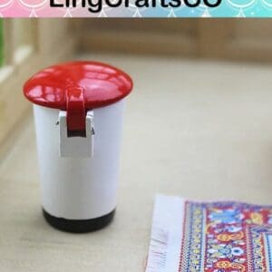 Miniature white trash can with red lid beside a colorful woven mat on a wooden surface.