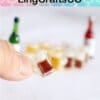 Close-up of a hand holding a tiny beer mug with blurred bottles in the background, labeled LingCraftsCO.