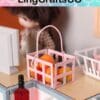 Miniature pink basket with toy fruits, wine bottle, and doll in background, on a dollhouse counter.