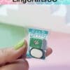 Hand holding a miniature model of soap in a small package with a colorful design.