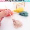 Hand holding a miniature yarn broom with others in different colors in the background, near a small chair.