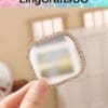 Hand holding a small, square rhinestone-framed mirror with a blurred interior background.