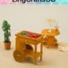 Miniature market cart with fruits and vegetables, beside a small table with glasses, plant, and toy animal on beige surface.