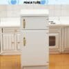 Miniature white refrigerator in a toy kitchen setup with cabinets and stove in the background.