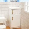 Miniature white cabinet with gold handles in a tiled room, next to a white chair and window.