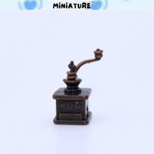 Miniature antique-style coffee grinder with "CAFÉ" engraved, set against a light background with heart motifs.