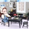 Doll in overalls and bunny hat sits at a miniature black table with books, flowers, in a detailed room setting.